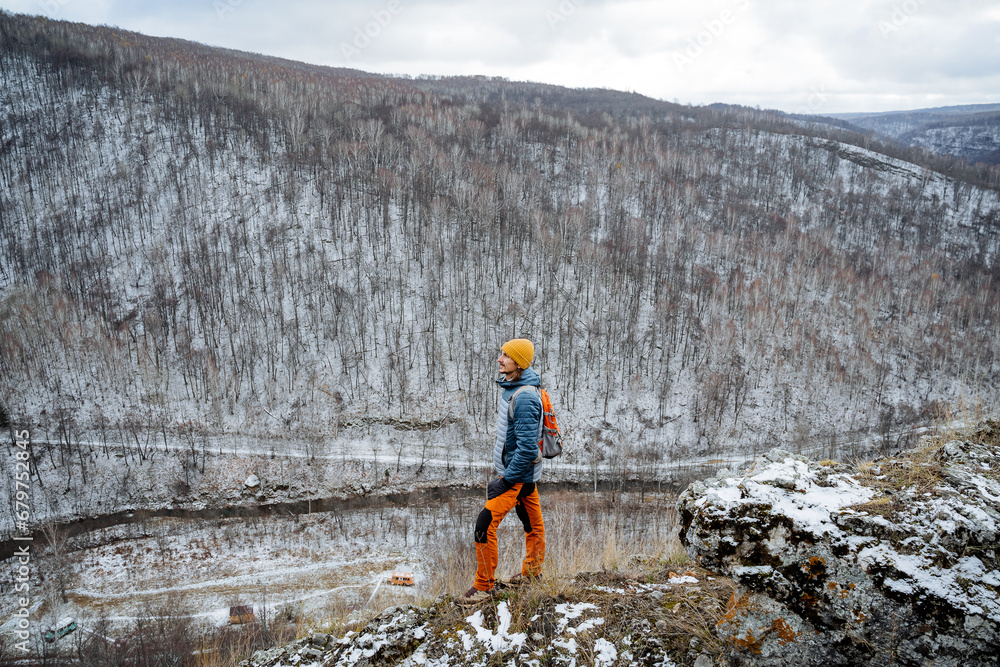Obraz premium Man standing on the edge of a cliff looking into the distance, traveling through the foothills, first snow in the mountains, bare forest, hiking alone in the mountains.