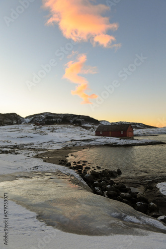 A traditional norwegian red barn enclosed by rugged mountains and snow-covered beaches in the arctic. Shown during the golden hour of a brief period of daylight near the town of Bodo.