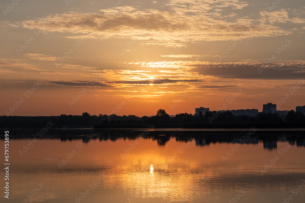 Scenic warm sunrise sun on cloudy sky reflecting in calm lake water. Peaceful morning in city park by the river