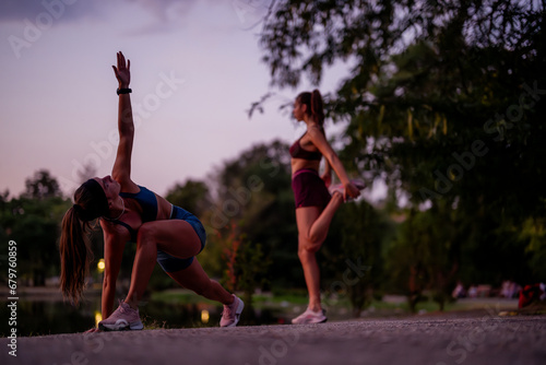 Fit girls exercising outdoors at night in a green park, stretching and warming up for a workout. Attractive athletes with muscular builds, training in a nature setting near a small lake.