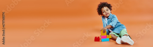 african american toddler boy in denim shirt sitting and playing building blocks on orange, banner photo