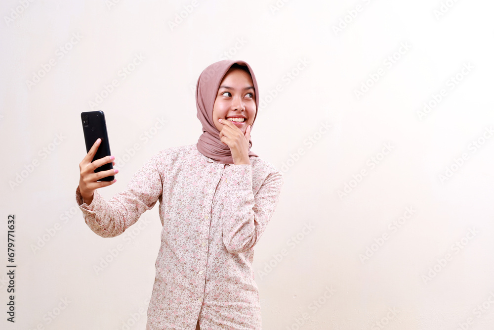 Happy young asian muslim girl holding a cell phone while looking at empty space beside her. Isolated on white background Happy young asian muslim girl holding a cell phone while looking at empty space beside her. Isolated on white background