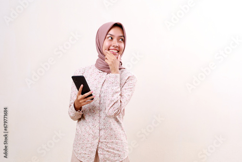 Happy young asian muslim girl holding a cell phone while looking at empty space beside her. Isolated on white background