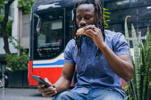 young black man with dreadlocks sitting using and checking phone and eating cookie outdoors.