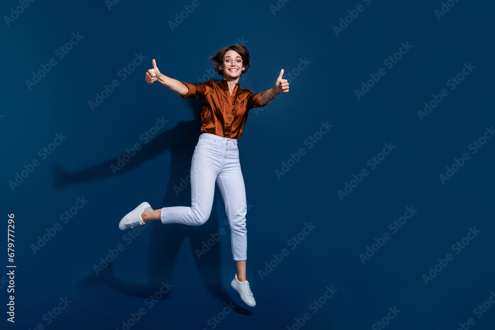 Full length photo of adorable confident lady wear brown shirt jumping showing two thumbs up empty space isolated blue color background