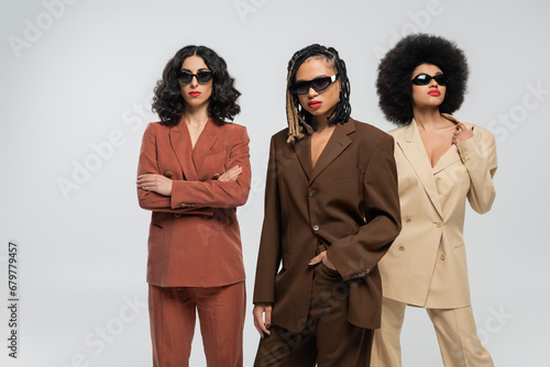 african american woman in sunglasses with hand in pocket near multiracial models in colorful suits
