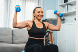 Athletic and sporty senior woman sitting on fit ball while engaging in weight lifting with dumbbell at home exercise as concept of healthy fit body lifestyle after retirement. Clout