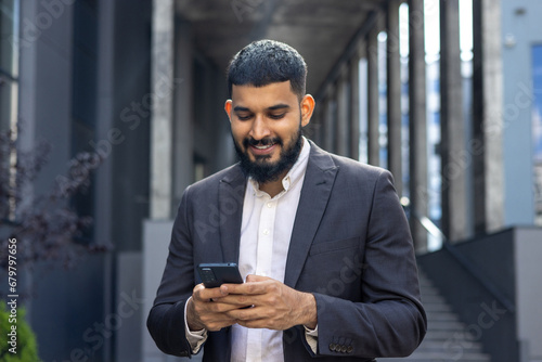Indian young male businessman standing on the street near an office building and using a mobile phone, dialing a number, writing a message, reading the news
