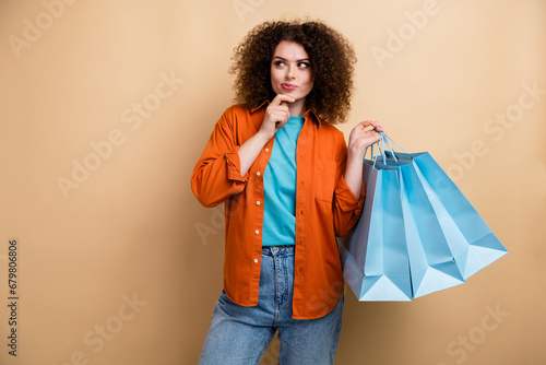 Photo of charming minded girl wear trendy clothes hold bags hand touch face chin kook empty space isolated on beige color background photo