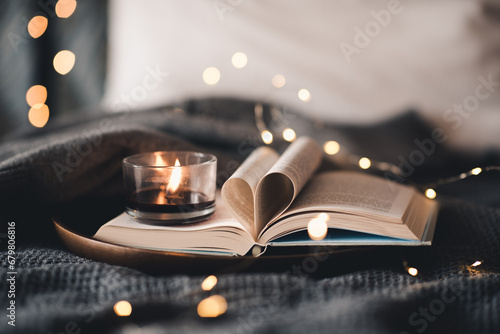 Open paper book with folded pages in heart shape and burning scented burning candle on tray in bed over Christmas lights close up. Winter holiday season. Valentines Day.