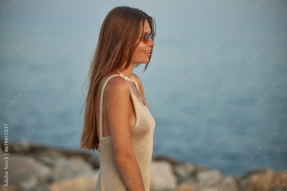 Cute stylish girl posing on the beach. The girl is dressed in a stylish off-the-shoulder dress. She's wearing brown sunglasses. The ocean is behind her. Cute stylish girl posing on the beach. The girl is dressed in a stylish off-the-shoulder dress. She's wearing brown sunglasses. The ocean is behind her.