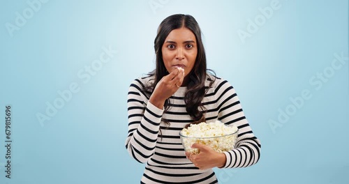 Popcorn, shock and woman in a studio for movie, film or show with entertainment or fun. Surprise, crazy and portrait of young Indian female person eating snack for cinema isolated by blue background. photo