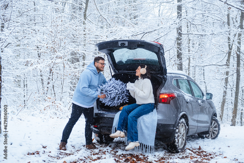 A woman sits with her cup in a car trunk in the woods, a man puts a Christmas tree there