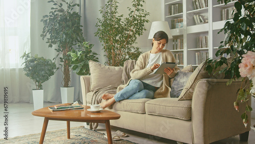 Young beautiful woman looking at her tablet in her apartment. Enjoying carefree peaceful weekend relaxation time alone in living room. Lady spending time at home with cell gadget technology photo