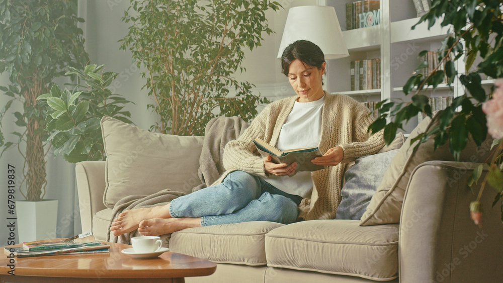 Portrait of beautiful woman relaxing, reading book while relaxing on cozy sofa at home. Smiles, enjoys leisure, carefree weekends with favorite literature and spending time. Habit, development, hobby