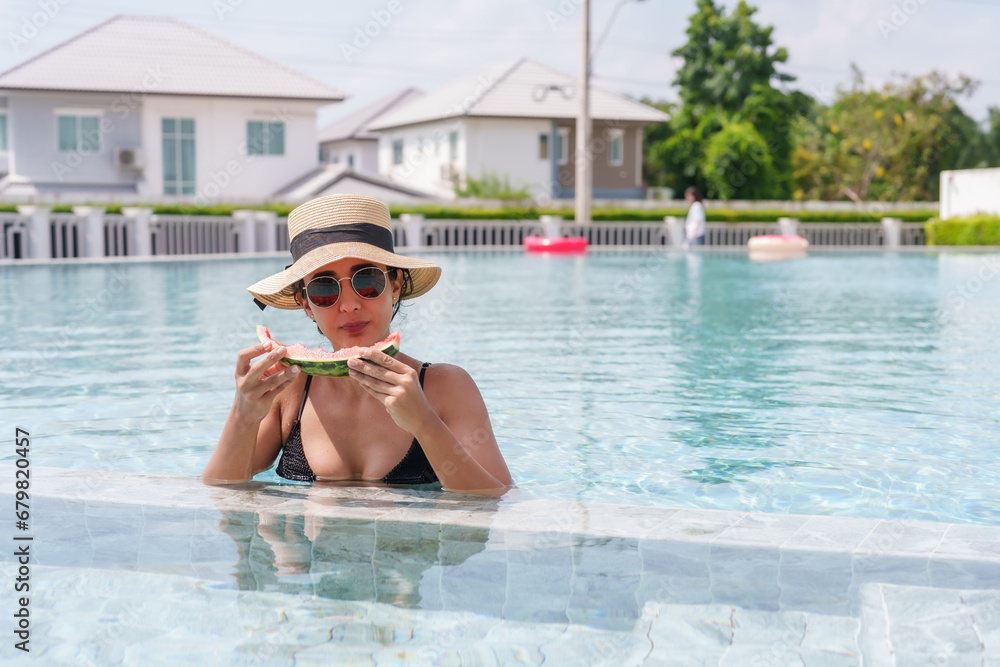 Beautiful Hispanic girl standing in water happily eating watermelon in pond Sunbathing in a swimsuit Wear a hat and sunglasses in the pool. On a clear summer day A relaxing afternoon on the weekend