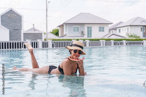 Beautiful Hispanic woman lying on edge swimming pool, wearing swimsuit sun hat sunglasses, woman lies down eats juicy red watermelon in comfort, body is wet because just finished swimming. © Ekkasit A Siam