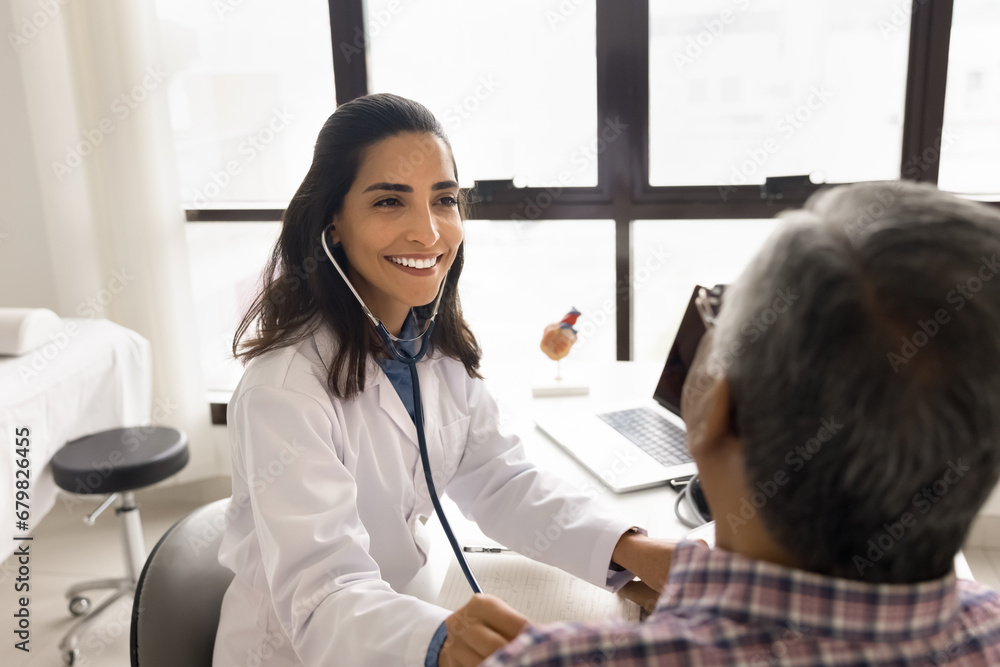 Fototapeta premium Positive beautiful young practitioner woman examining senior patient man, applying stethoscope to chest, checking heartbeat rate, breathing, lungs work, sitting at workplace table