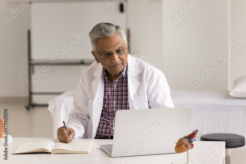 Focused senior Indian doctor man making records in medical history book at laptop, using computer, giving online consultation, looking at display, sitting at workplace in practitioner office