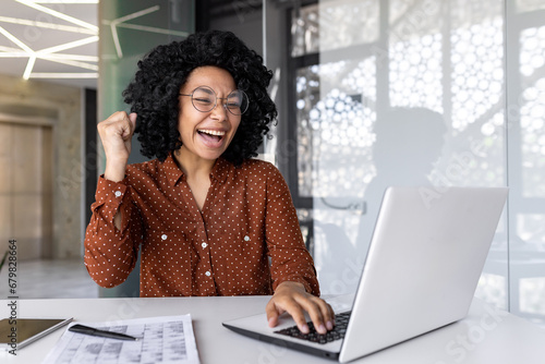 Φωτογραφία Joyful successful satisfied african american businesswoman celebrating great achievement results at workplace inside office, female worker celebrating holding hand up smiling with laptop