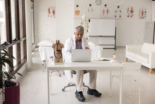 Senior Indian doctor man in white coat typing on laptop in clinic, sitting at workplace with body organs model on table, working in hospital office interior with medical education posters photo
