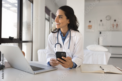 Happy young doctor woman using online medical application on smartphone, holding mobile phone at workplace, giving consultation on Internet, looking away, thinking, enjoying modern technology photo