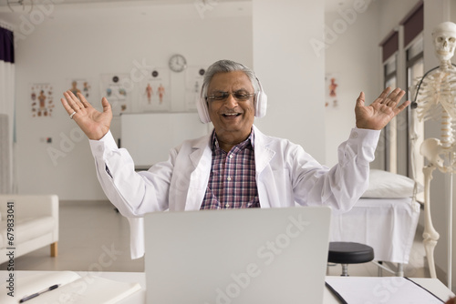 Cheerful elderly Indian doctor chatting with patient on video call, speaking at laptop, laughing, having fun, opening hands with joy, getting good news about treatment success