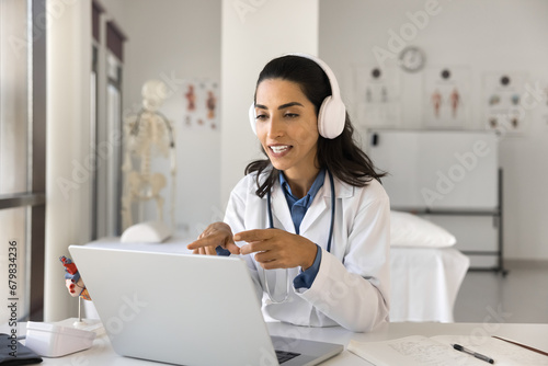 Young Latin doctor woman in wireless headphones speaking on video conference call at laptop, talking to patient online in hospital office, giving lecture, webinar to medical students photo