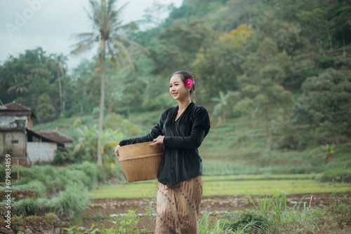 Young woman from an Indonesian village carrying a basket or basket made of woven bamboo. Asian village woman standing and walking in the village rice fields.