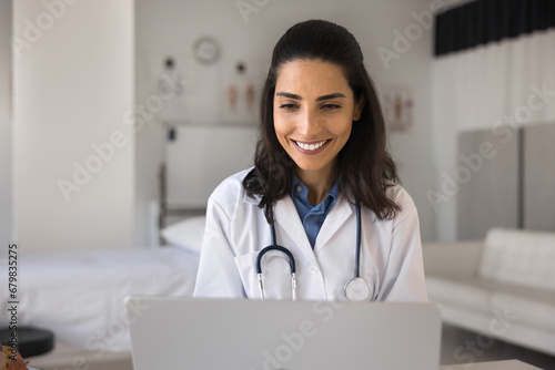 Cheerful pretty young Latin doctor woman enjoying work at laptop in surgery office, smiling, laughing, talking on video call, looking at screen, getting good news giving consultation on Internet