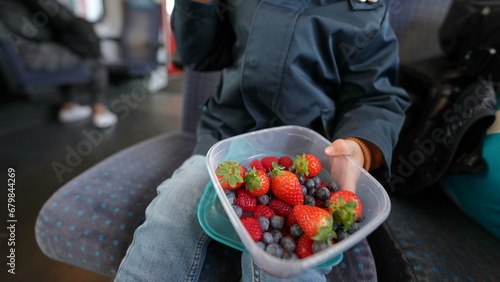 Fotografie Child holding berries inside travel container eating healthy fruit snack while s