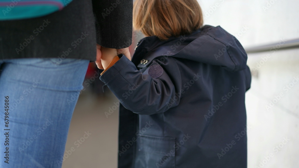 Back of small boy holding hands with mother walking while on commute travel, child wearing jacket