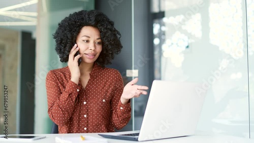 Busy young african american female employee talking on smartphone while sitting at workplace in business office. Focused black woman discusses business with a partner or has a conversation with client photo