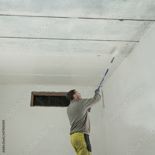 Diligent Worker in Grey Jumper and Yellow Trousers Adds Final Strokes to White Garage Paint Job.