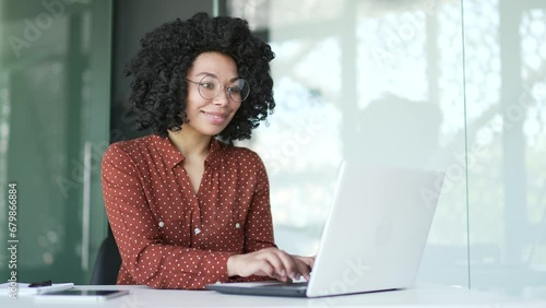 Young african american female employee typing on laptop sitting at workplace in business office. A smiling black woman in glasses works on a computer on a project, chats online, writes email messages photo
