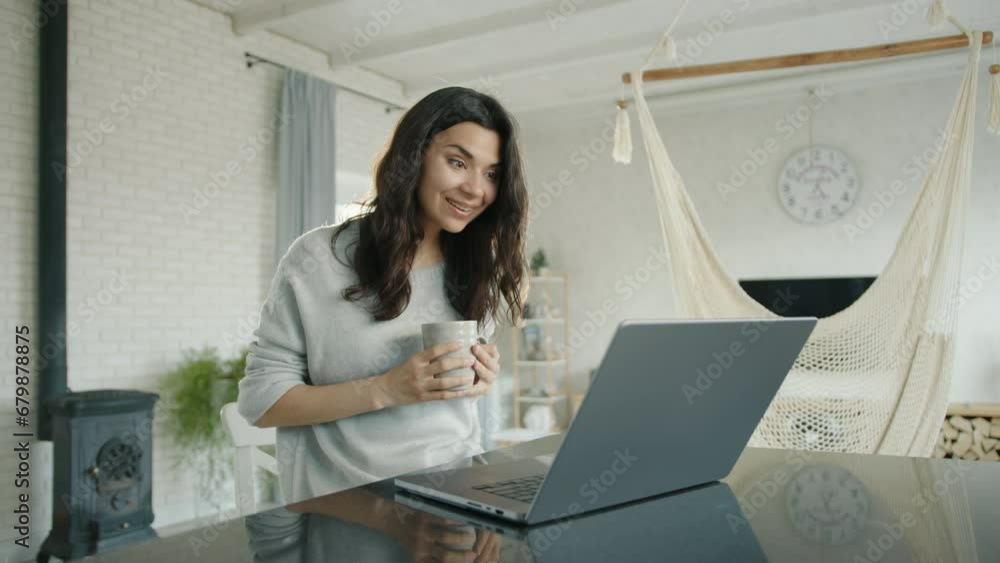 Pretty smiling brunette communicates via video link on laptop sitting at table. Young woman holds a cup, drinks, waves hello, has nice conversation. High quality 4k footage Pretty smiling brunette communicates via video link on laptop sitting at table. Young woman holds a cup, drinks, waves hello, has nice conversation. High quality 4k footage