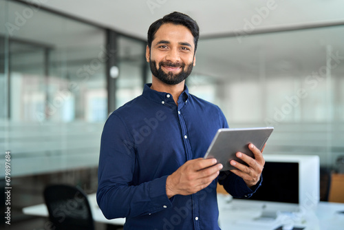 Smiling professional Indian businessman using tab computer working standing in office. Confident professional business man entrepreneur holding digital tablet tech device working, looking at camera. photo