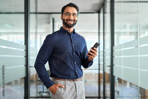 Happy Indian business man holding mobile cell phone device looking at camera standing at work. Smiling businessman executive using finance banking apps smartphone working on cellphone in office. photo
