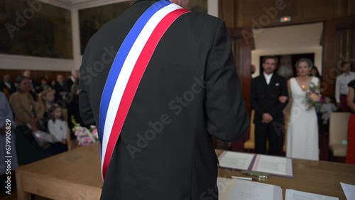 A mayor from behind with a French tricolor scarf making a speech to celebrate a heterosexual marriage in a town hall. Blurred background on the bride and groom. French wedding with two happy lovers.