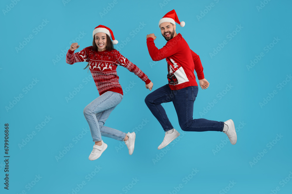 Happy young couple in Christmas sweaters and Santa hats jumping on light blue background