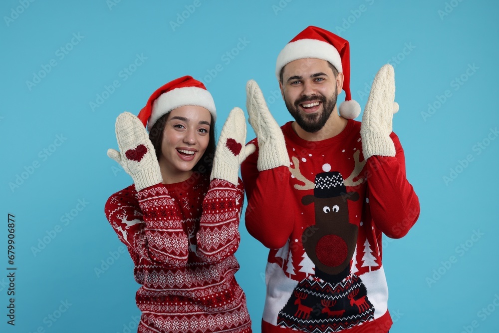 Young couple in Christmas sweaters, Santa hats and knitted mittens on light blue background Young couple in Christmas sweaters, Santa hats and knitted mittens on light blue background