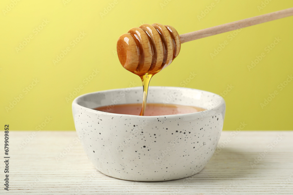Pouring tasty honey from dipper into bowl at white wooden table against yellow background, closeup Pouring tasty honey from dipper into bowl at white wooden table against yellow background, closeup