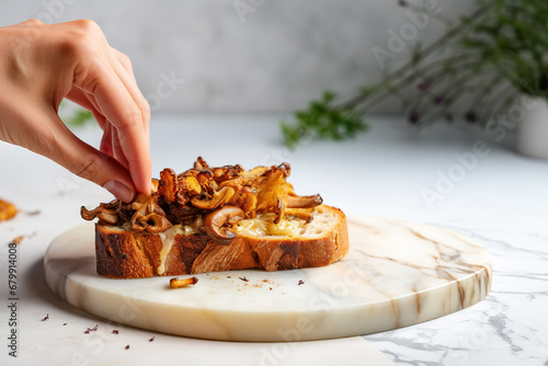Female hand making a toast with roasted chanterelles and cheese, on marble kitchen countertop. Fried wild mushrooms on toasted slice of bread.