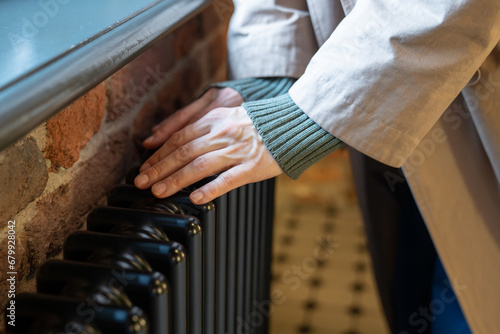 Cropped photo of woman warming hands near radiator at home, female touching old retro cast iron radiator in winter during heating season, checking heating system working capacity