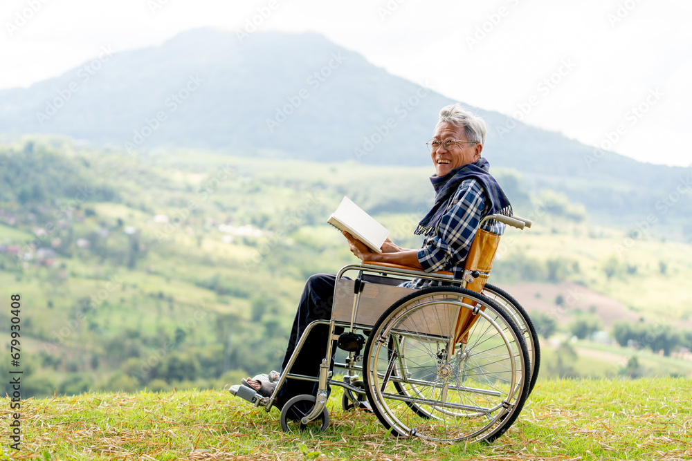 Asian senior man with eyeglasses hold the book and sit on wheelchair also look at camera and stay near the cliff with grassfield and mountain as background.