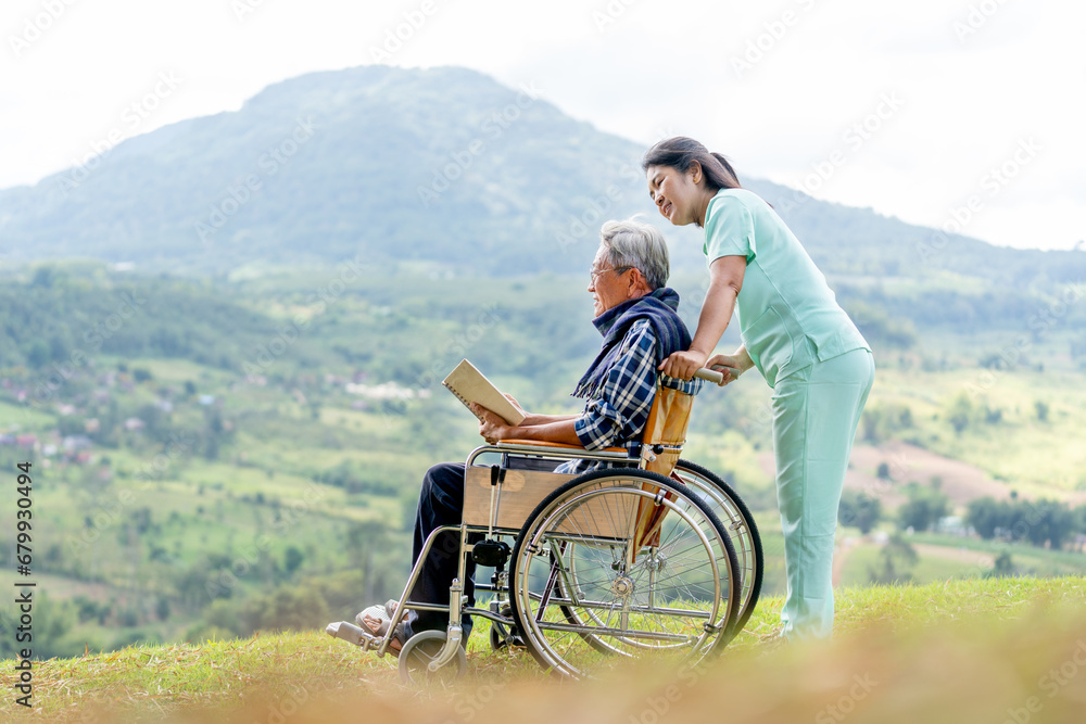 Asian nurse stand behide and hold wheelchair handle with senior man sit on wheelchair also hold the book and stay near cliff.