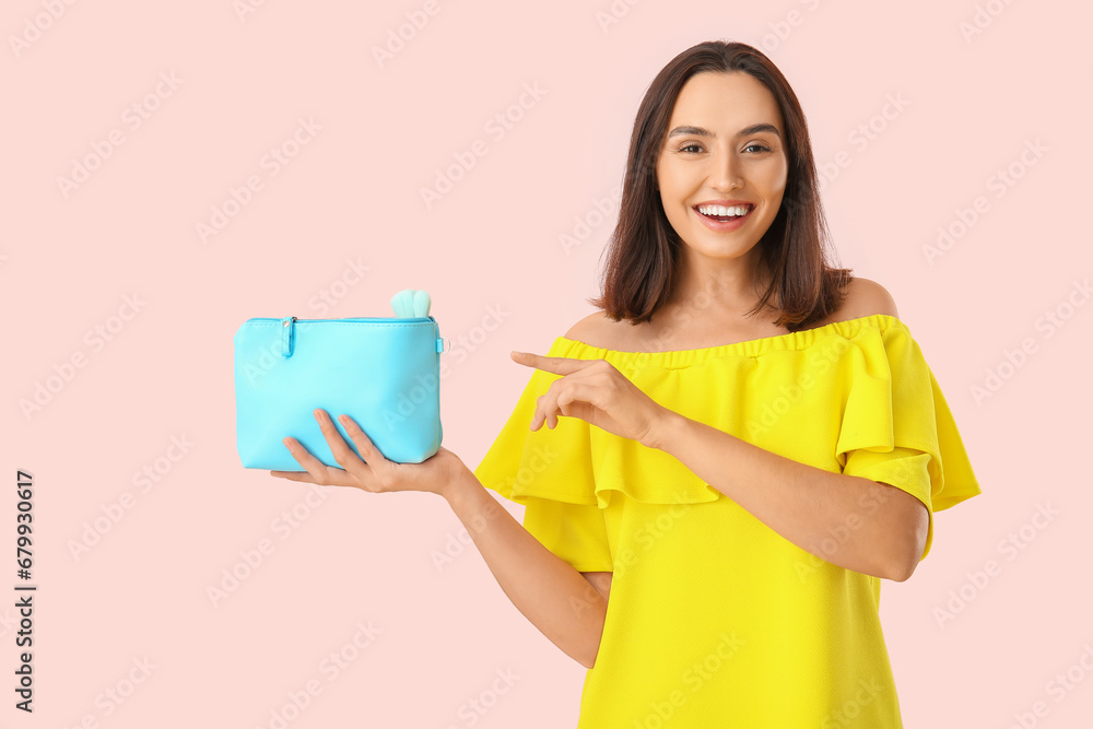 Beautiful young woman pointing at cosmetic bag full of makeup products on pink background Beautiful young woman pointing at cosmetic bag full of makeup products on pink background