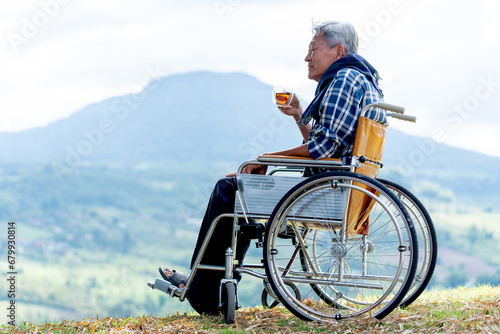 Asian senior man hold a cup of tea and sit on wheelchair with lood forward and stay near the cliff and mountain and grassfield as background.