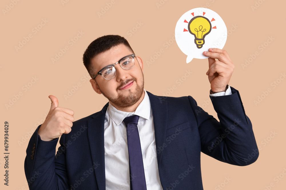 Young businessman holding speech bubble with drawn light bulb and showing thumb-up gesture on beige background. Business idea concept Young businessman holding speech bubble with drawn light bulb and showing thumb-up gesture on beige background. Business idea concept
