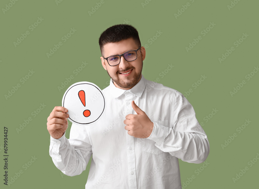 Young businessman holding sheet with exclamation mark and showing thumb-up gesture on green background. Business idea concept Young businessman holding sheet with exclamation mark and showing thumb-up gesture on green background. Business idea concept